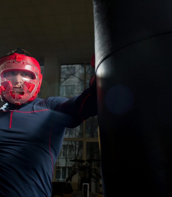 Young athlete in boxing helmet and gloves kicking punching bag in gym